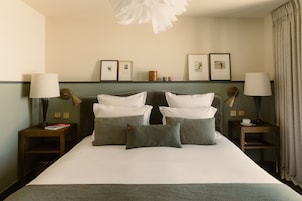 Una habitación de hotel con una cama grande, dos mesitas de noche con lámparas, un estante con marcos y un colgante de techo.