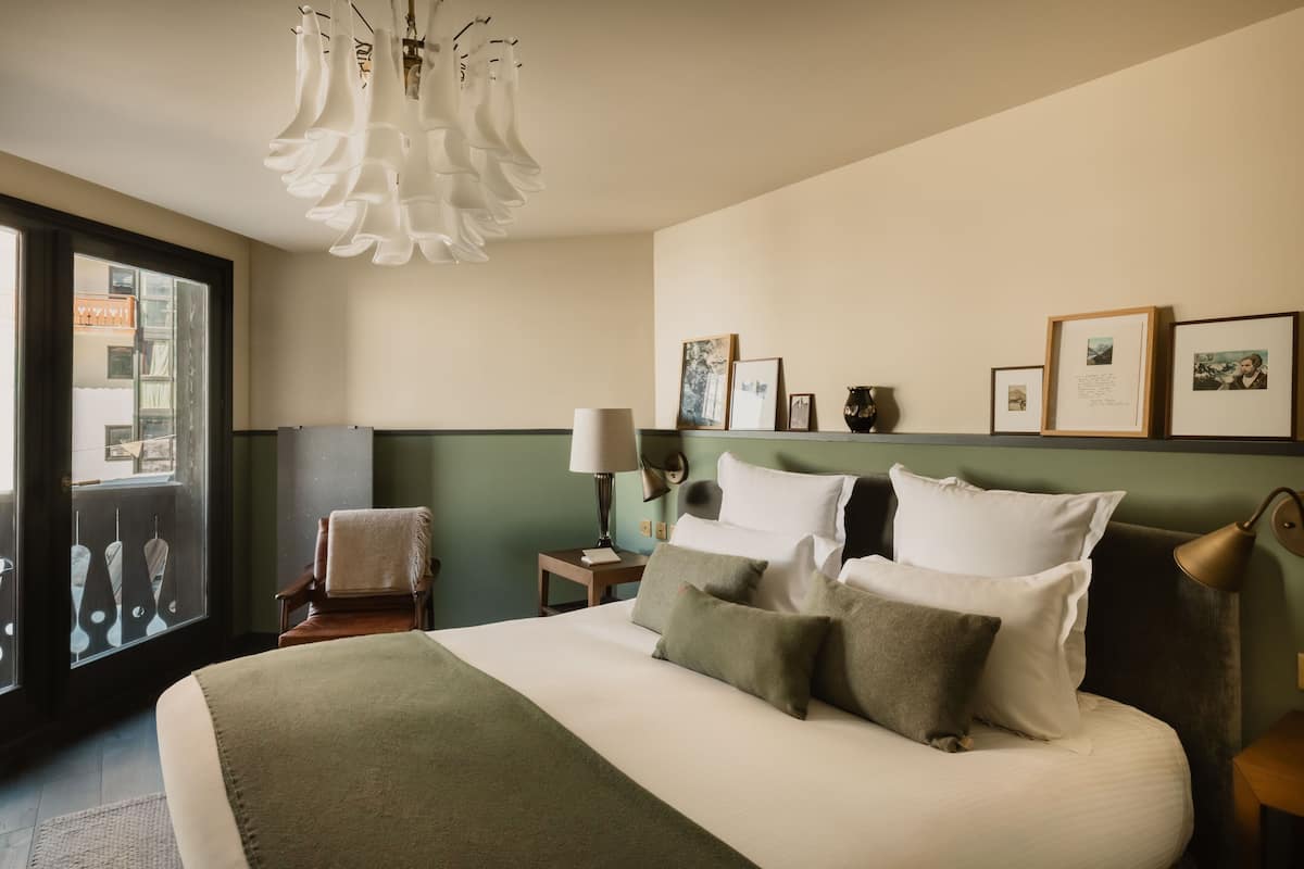 A hotel room with a large bed, a chair, a desk, a lamp, and framed pictures on the wall.