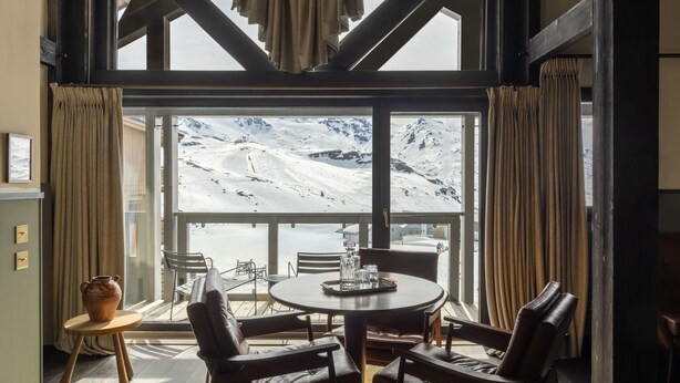 A room with a large window overlooking a snowy mountain, a round table with chairs, and a vase on a small table. - Le Fitz Roy (Les Belleville)