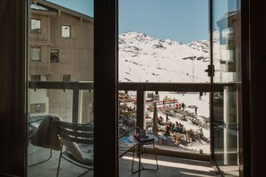 A view from a window with a chair and table inside, looking out to a snowy mountain and a busy outdoor area with people and structures. - Le Fitz Roy (Les Belleville)