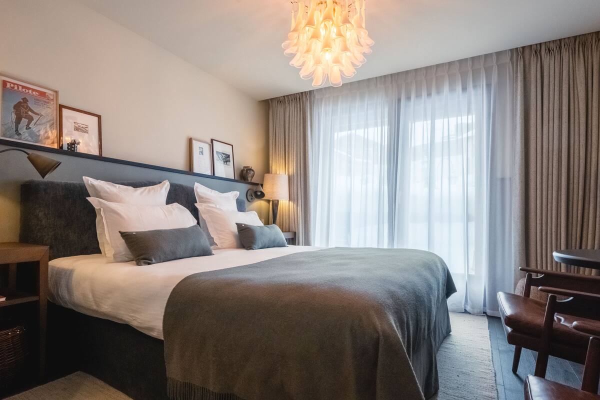 A hotel room with a bed, a desk, a chair, a lamp, and a large window with curtains.