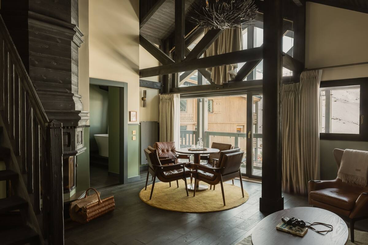 A spacious room with a high ceiling, a wooden staircase, a round table with chairs, a leather armchair, and large windows.
