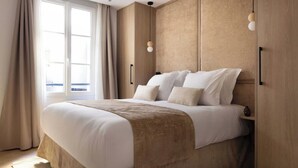 Een groot bed met een beige hoofdeinde en een bruine plaids, een raam met gordijnen, en een hanglamp aan de muur.
