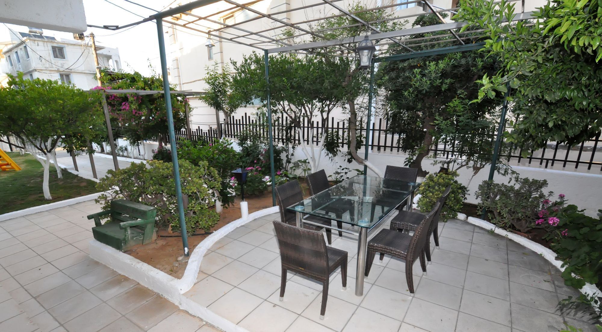 An outdoor patio with a glass table and chairs, surrounded by greenery and a white fence.