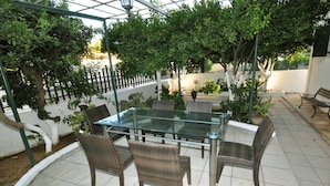Un patio al aire libre con una mesa de vidrio y sillas de mimbre, rodeado de macetas con plantas y una cerca blanca.