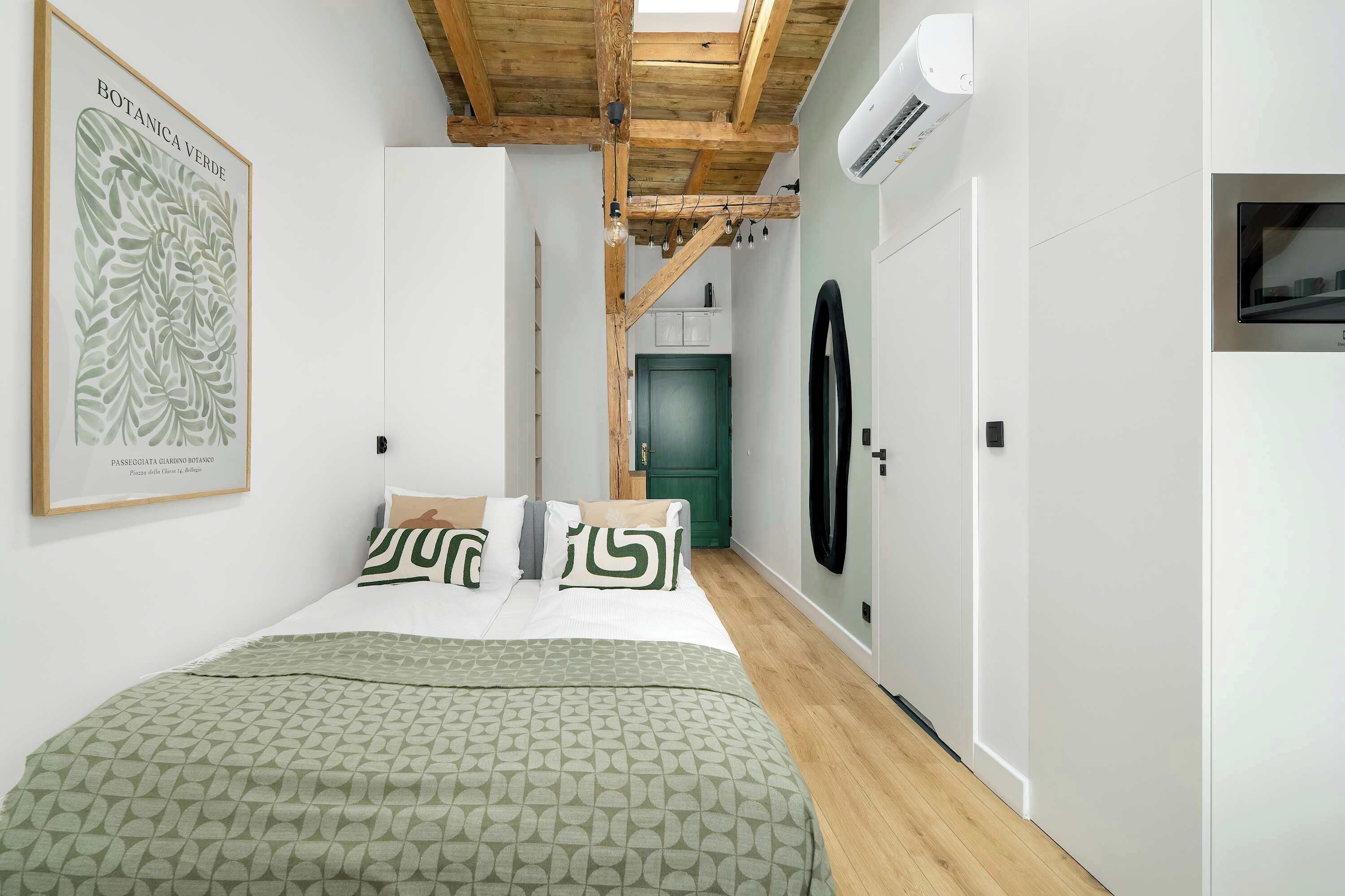 A bedroom with a bed, a green patterned bedspread, a framed botanical print on the wall, and a wooden ceiling.