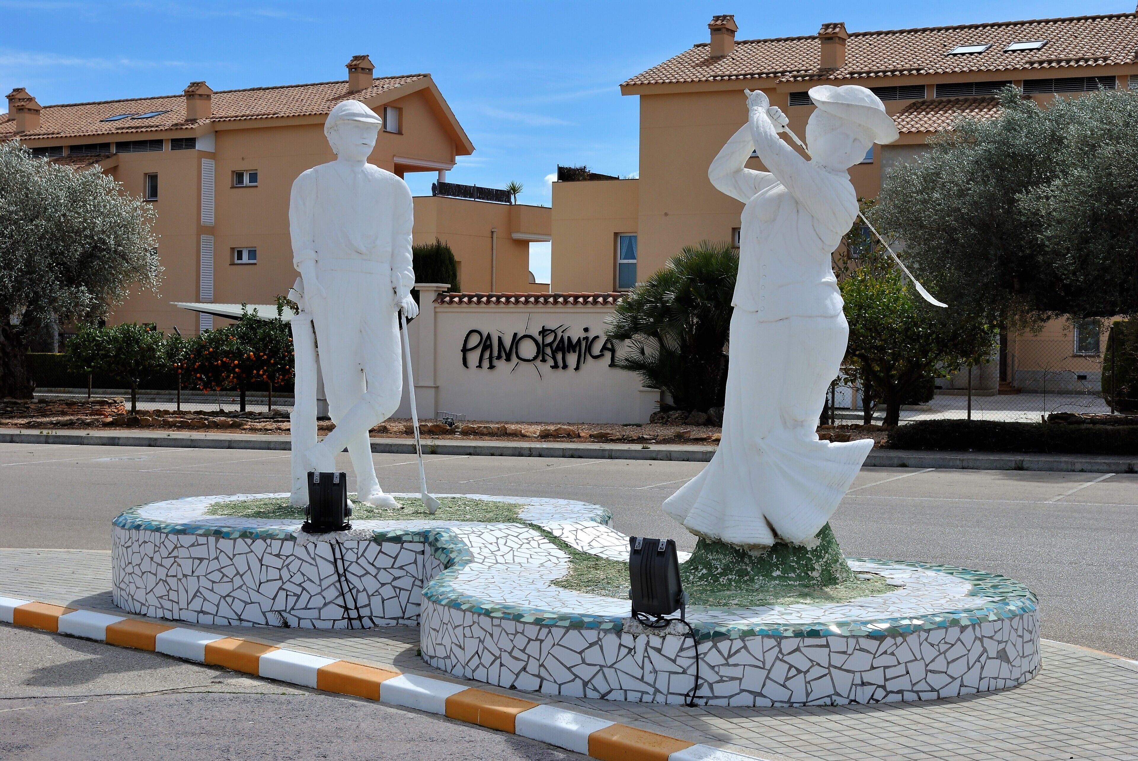 Dos estatuas blancas sobre plataformas circulares con mosaicos, ubicadas en una plaza pública con un edificio al fondo.