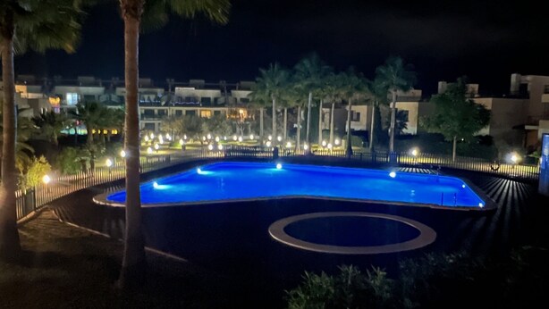Ein Schwimmbecken im Dunkeln mit kreisförmigem Design, umgeben von Palmen und einem Zaun, mit Gebäuden im Hintergrund.