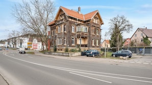 A large, historic building with a red-tiled roof and multiple windows, located beside a road with parked cars and a pedestrian crossing. - Fully equipped apartments in Metzingen - ideal for fitters & companies (3 units) (Metzingen)