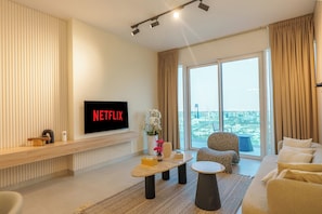Un salon moderne doté d’un téléviseur à écran plat affichant le logo « NETFLIX », d’un canapé, d’une table basse et offrant une vue sur le paysage urbain à travers de grandes fenêtres.