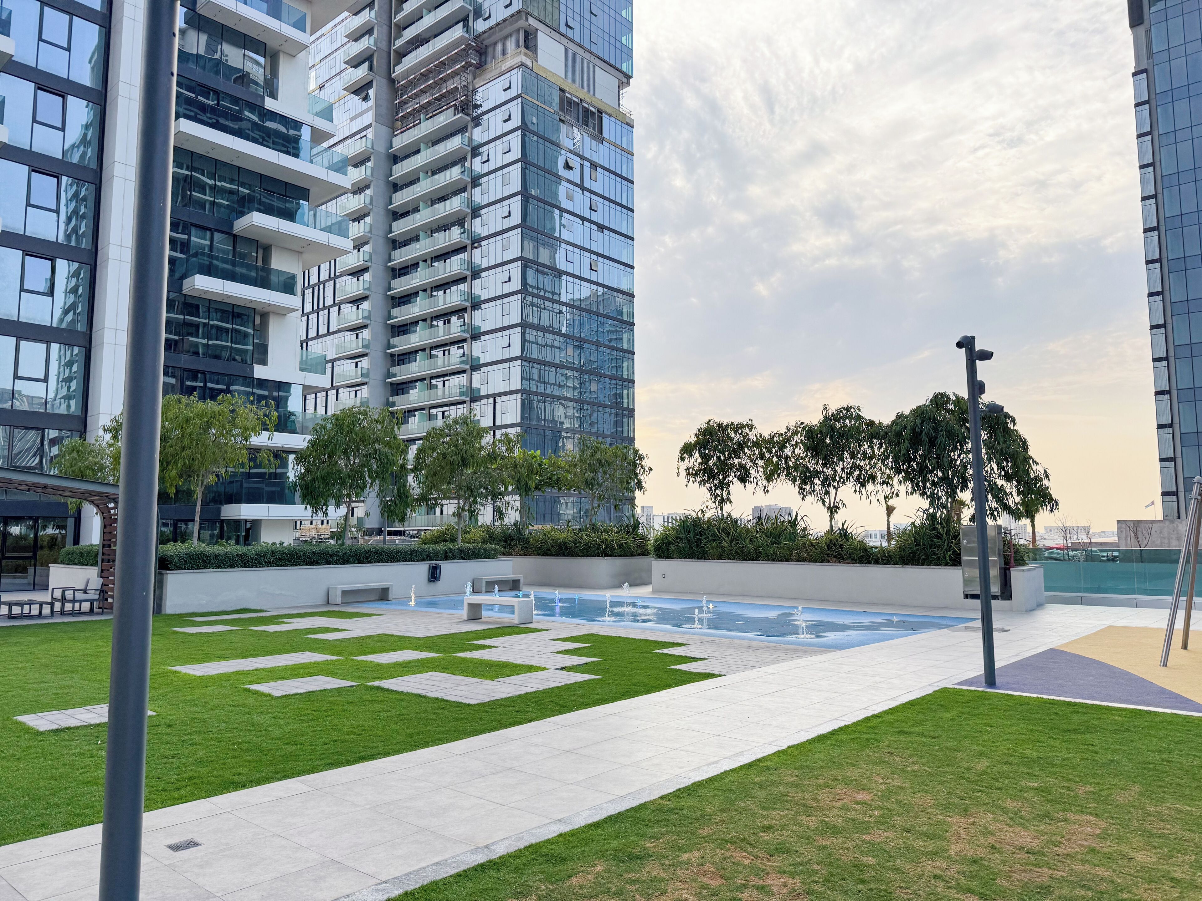 A modern urban setting with a large building, a landscaped area with grass and pathways, and a water feature.