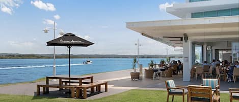 A waterfront restaurant with outdoor seating, a view of the water, and a modern building in the background.