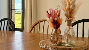 A wooden dining table with two glass vases containing dried flowers, a framed picture on the wall, and a view of a grassy field through a window. - Lovely Holiday Cottage in Corseul (Corseul)