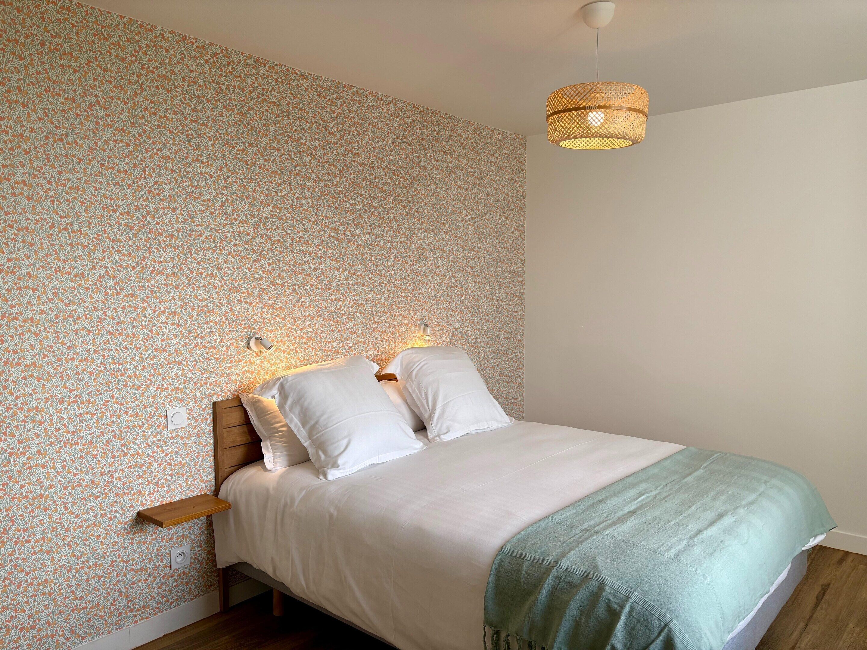 Ein Schlafzimmer mit strukturiertem Wandpapier, einem Bett mit weißer Bettwäsche, einer grünen Tagesdecke und einer hängenden Leuchte.