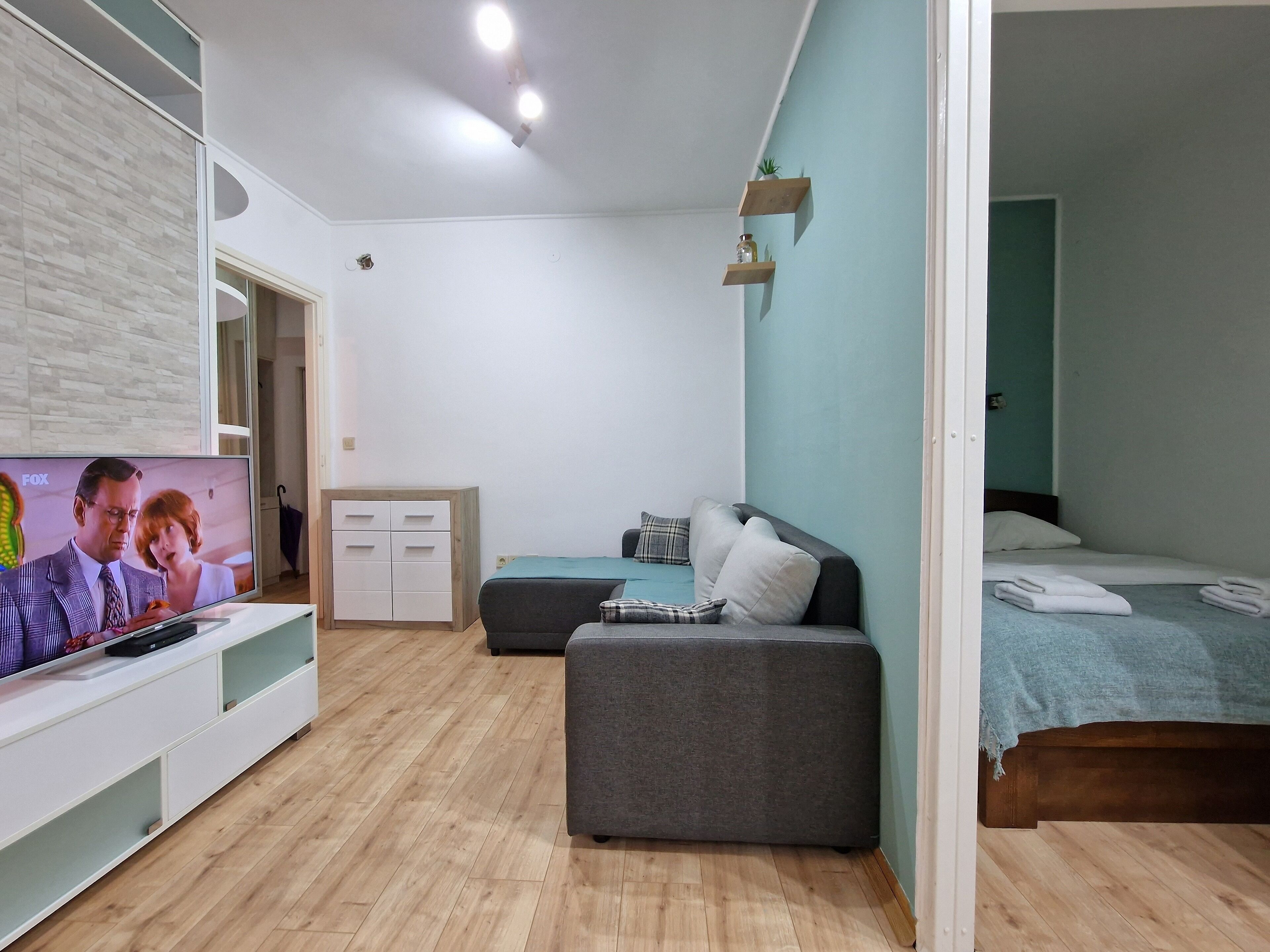 Um quarto moderno com um sofá cinza, uma cama com colcha azul, uma cômoda de madeira e uma TV exibindo uma cena de filme.