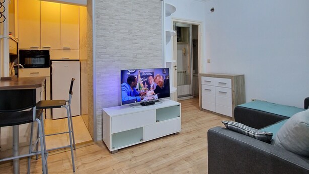 Ein modernes Wohnzimmer mit Flachbildfernseher, einer Couch, einem Essbereich mit Barhocker und einer Küche mit weißen Schränken.