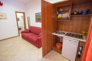 A compact kitchen with a sink, stove, and storage shelves. A red sofa is in the adjacent living area. - Hortensia House (Cefalù)