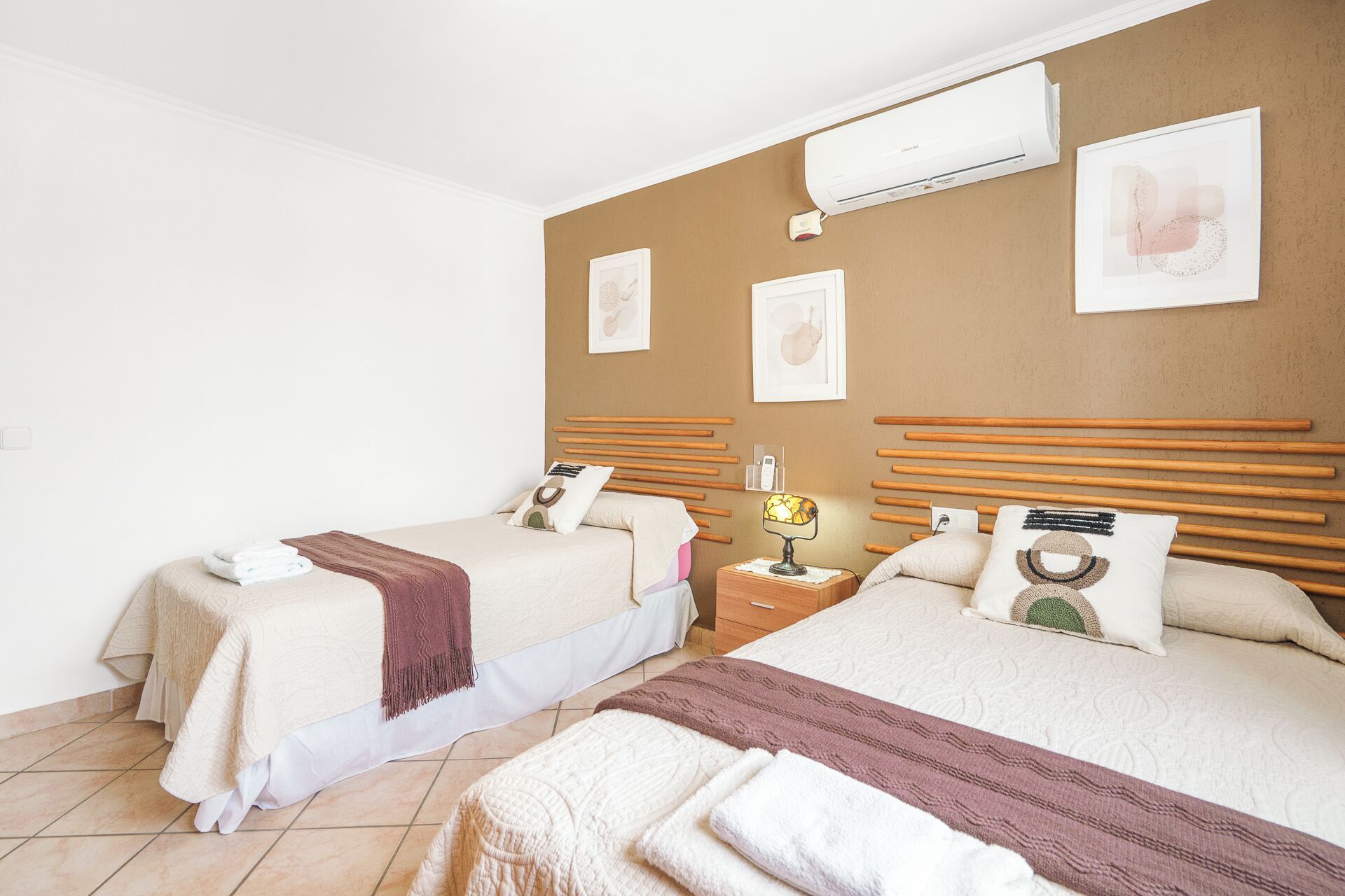 A hotel room with two beds, a nightstand, a wall-mounted air conditioner, and framed artwork on the wall.