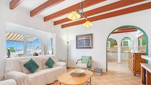 A spacious living area with a white sofa, a round wooden table, and a view of the kitchen through an archway. - Private villa with barbecue, ping pong, swimming pool and garden. 7 min from the beach! (Cala en Porter)