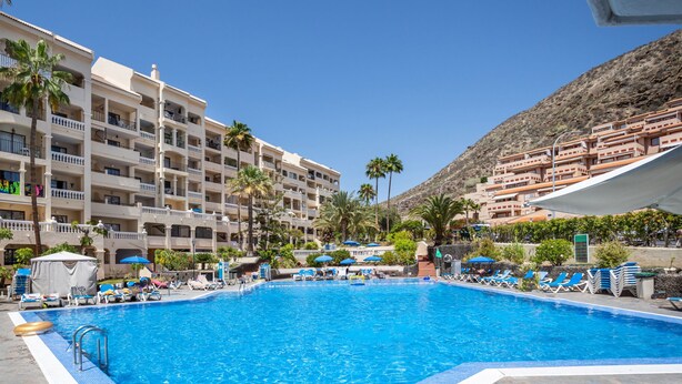A swimming pool with lounge chairs and umbrellas, surrounded by multi-story buildings with balconies and palm trees. - Central Studio Apartment. Communal Heated Pool (CN)