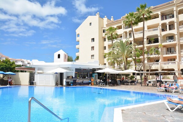 Une piscine à l’eau d’un bleu limpide, entourée de chaises longues et de parasols, avec un bâtiment visible au loin.