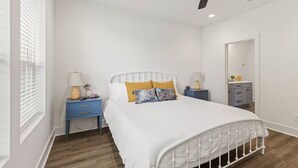 Ein Schlafzimmer mit einem weißen Bett, zwei Nachttischen, einem Deckenventilator, einem Fenster mit Jalousien und einem Spiegel.