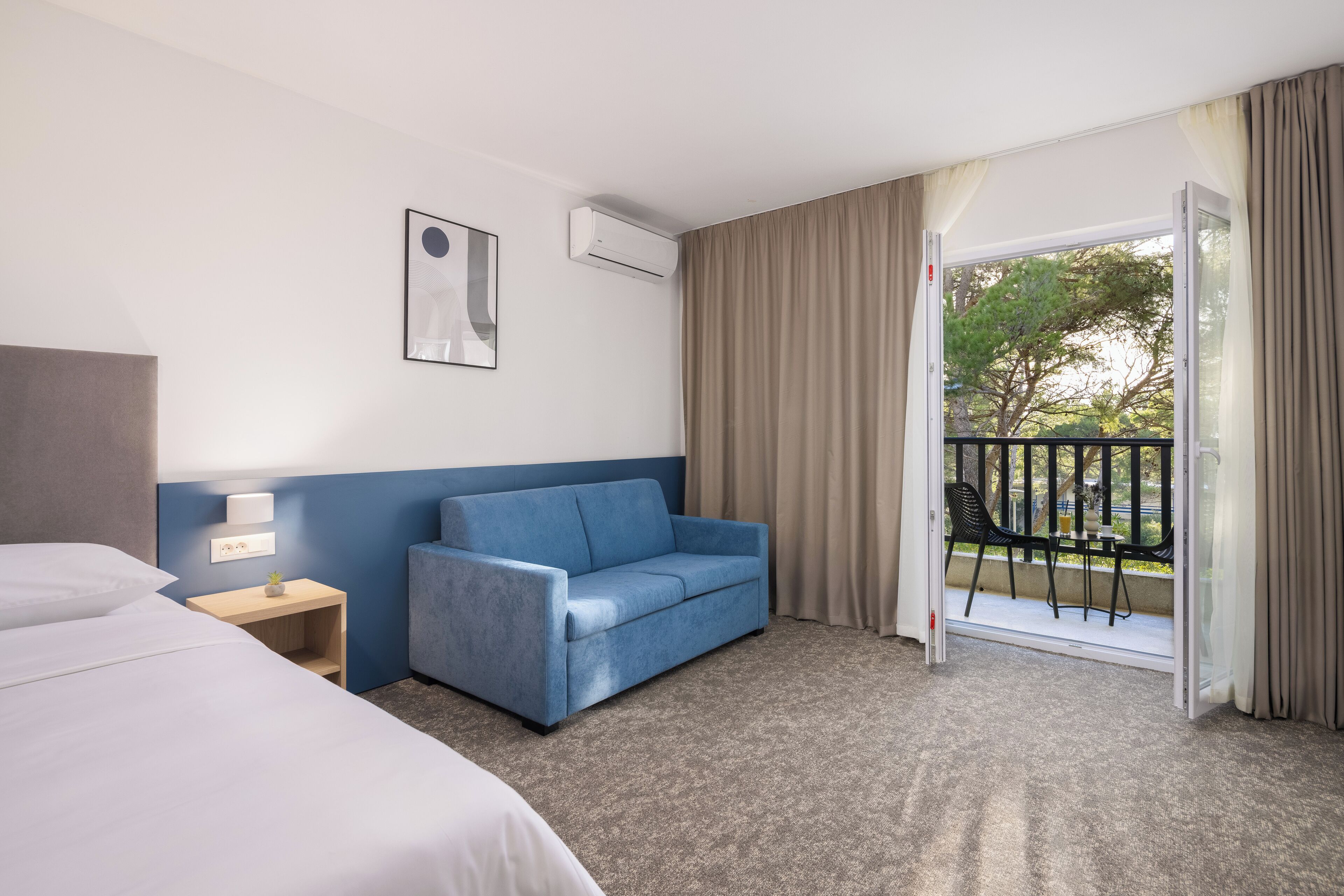 A hotel room with a bed, a blue sofa, a small table with a lamp, and a balcony with a view of trees.