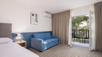 A hotel room with a bed, a blue sofa, a small table with a lamp, and a balcony with a view of trees.