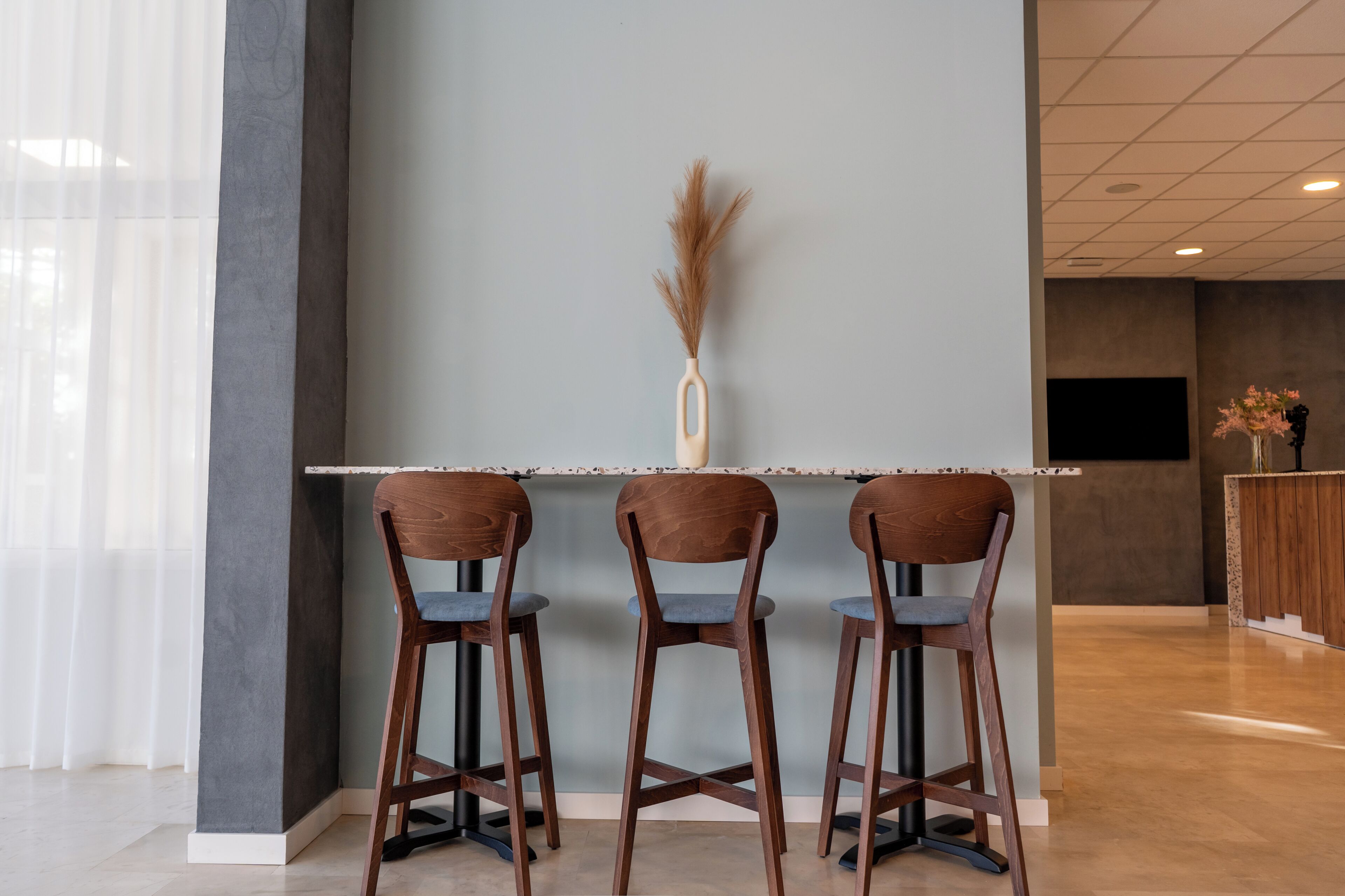 Un espace bar avec trois tabourets de bar en bois, un vase central contenant des plantes séchées et un aménagement intérieur moderne.