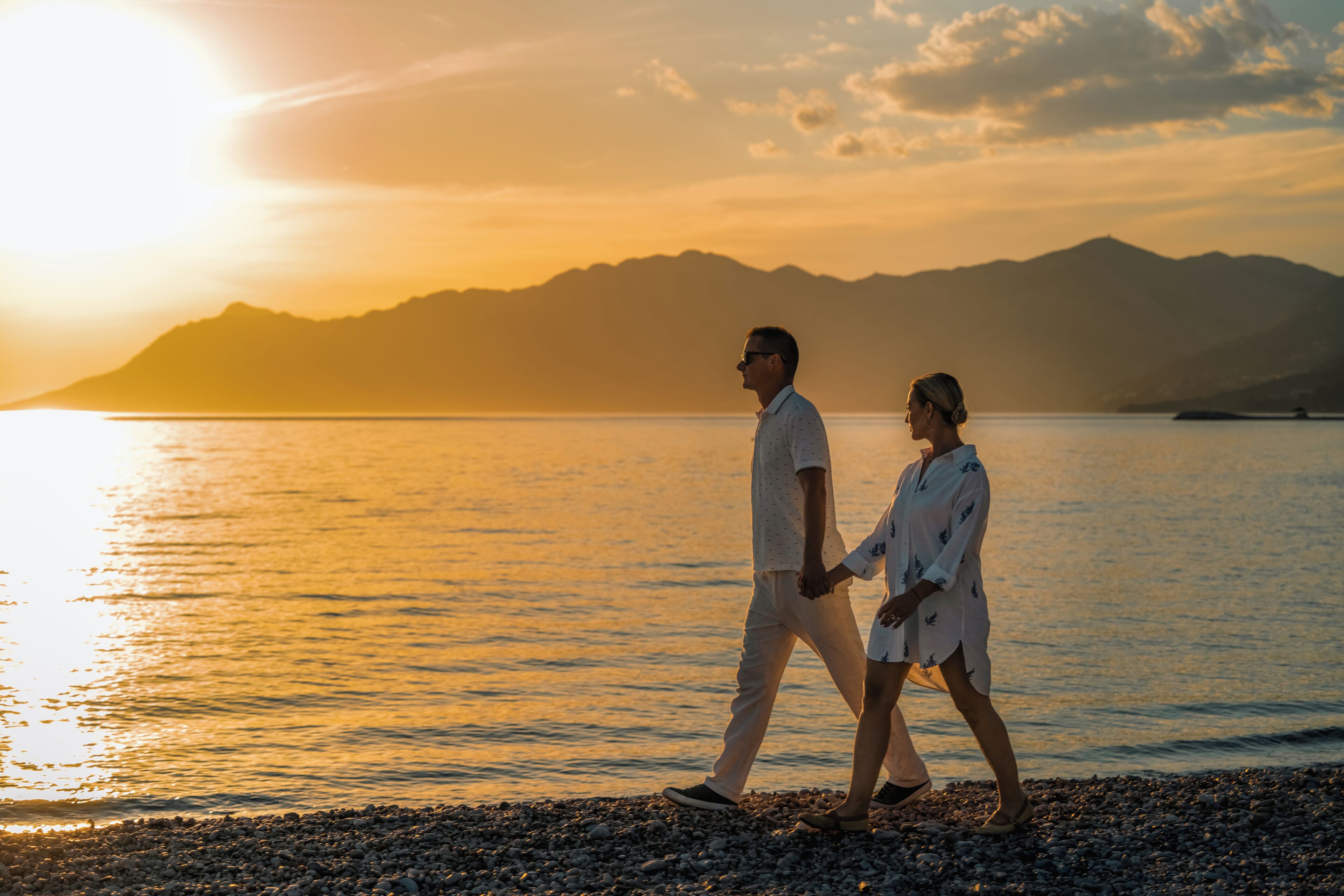Una pareja caminando sobre una playa de piedrecitas con el atardecer de fondo.