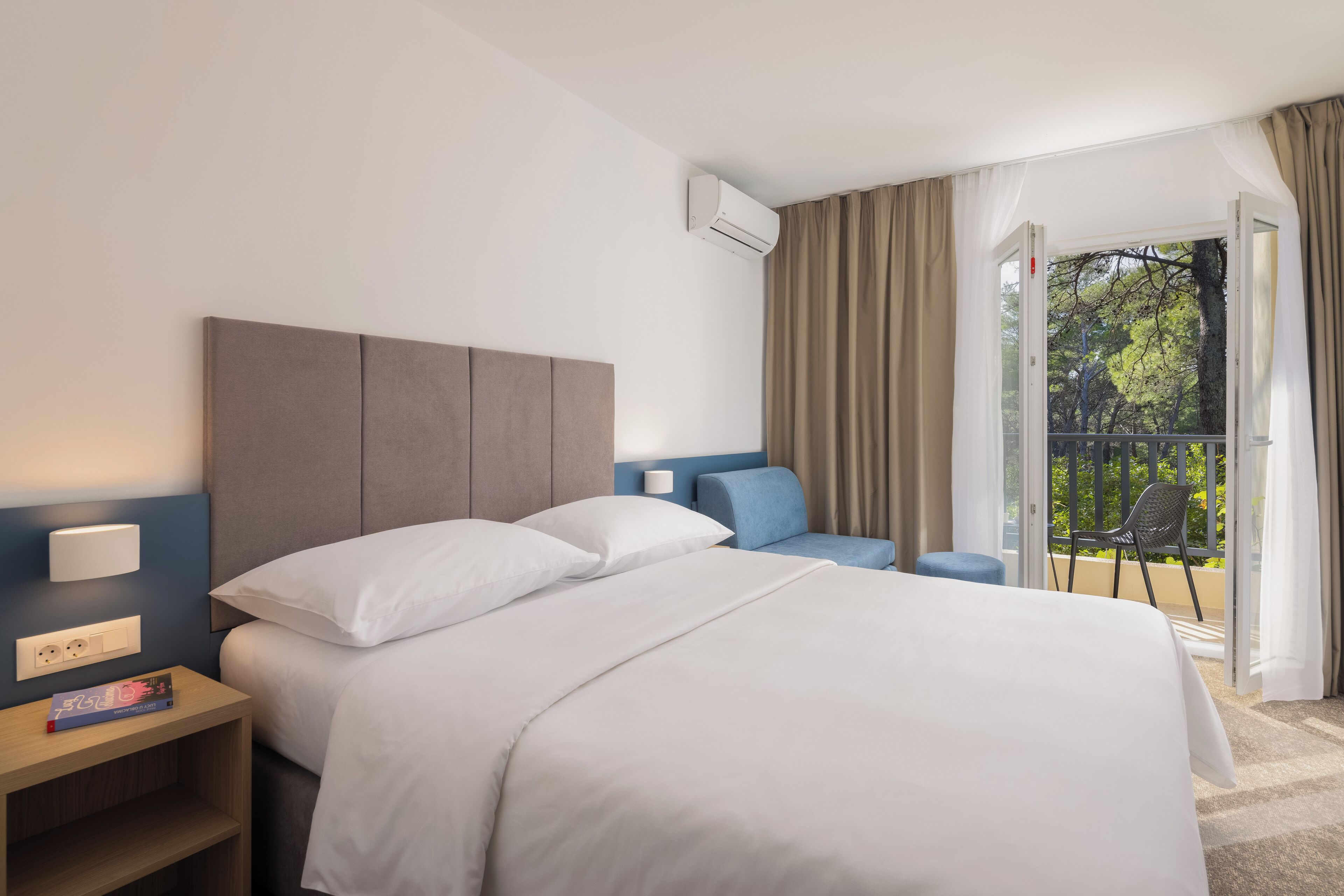 A hotel room with a bed, bedside table, lamp, and a view of the outdoors through a sliding glass door.