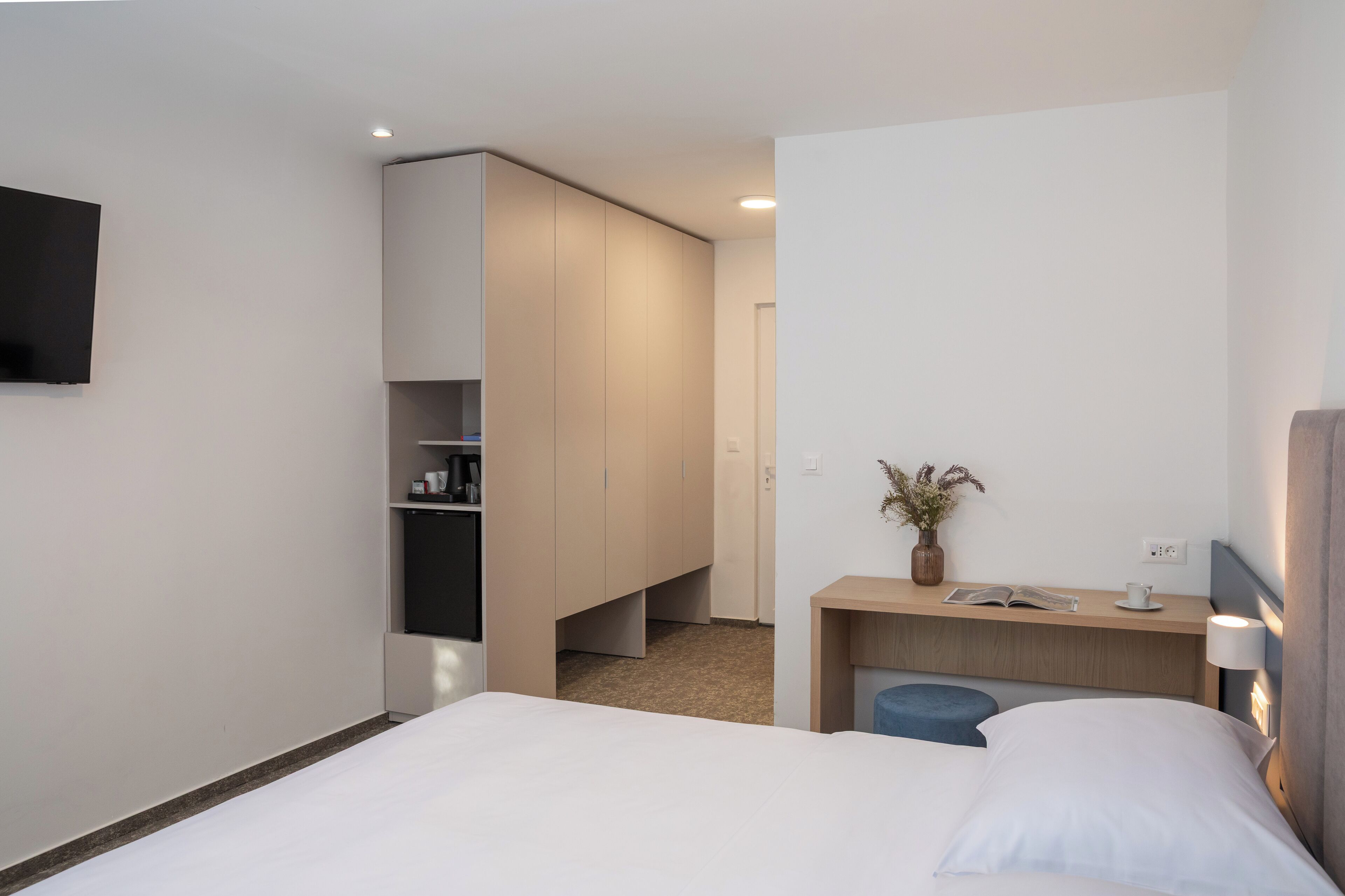 A modern hotel room with a bed, a desk with a lamp, a small seating area, and a wardrobe.