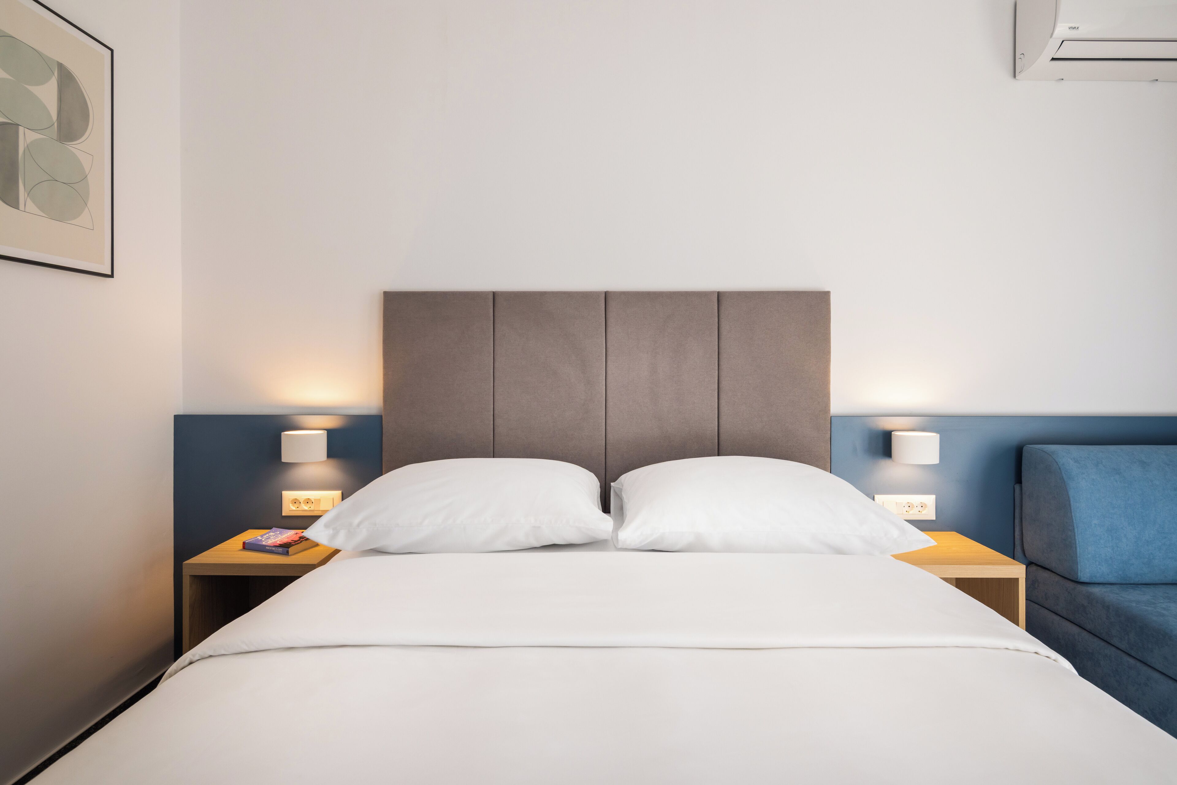 A hotel room with a bed, two bedside tables, a nightstand, a blue armchair, and a wall-mounted air conditioner.