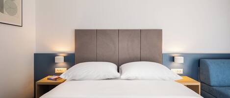 A hotel room with a bed, two bedside tables, a nightstand, a blue armchair, and a wall-mounted air conditioner.