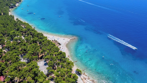 An aerial view of a coastal area with a beach, clear blue waters, and dense green forest. - Alemaris Resort (Baška Voda)