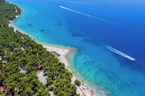 An aerial view of a coastal area with a beach, clear blue waters, and dense green forest. - Alemaris Resort (Baška Voda)