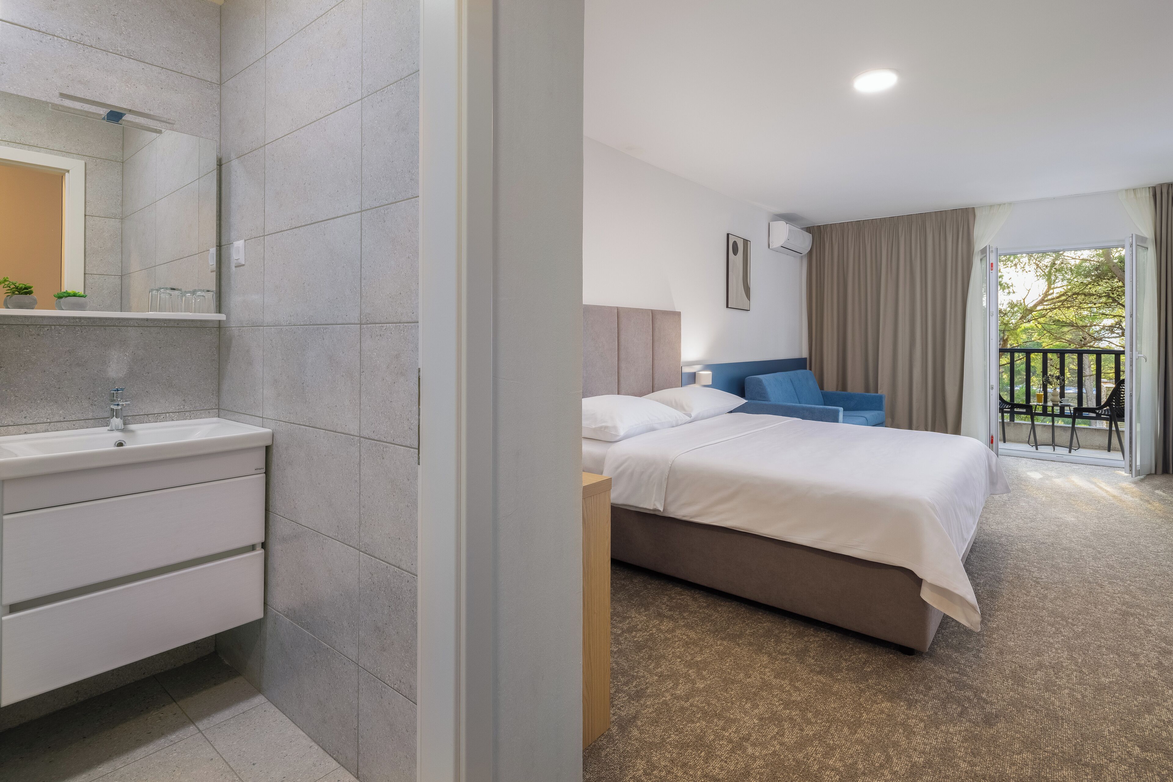 A modern hotel room with a bathroom, a bed with white bedding, a blue headboard, and a balcony with a view.