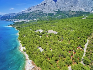 An aerial view of a coastal area with dense greenery, a beach, and a mountainous backdrop. - Alemaris Resort (Baška Voda)