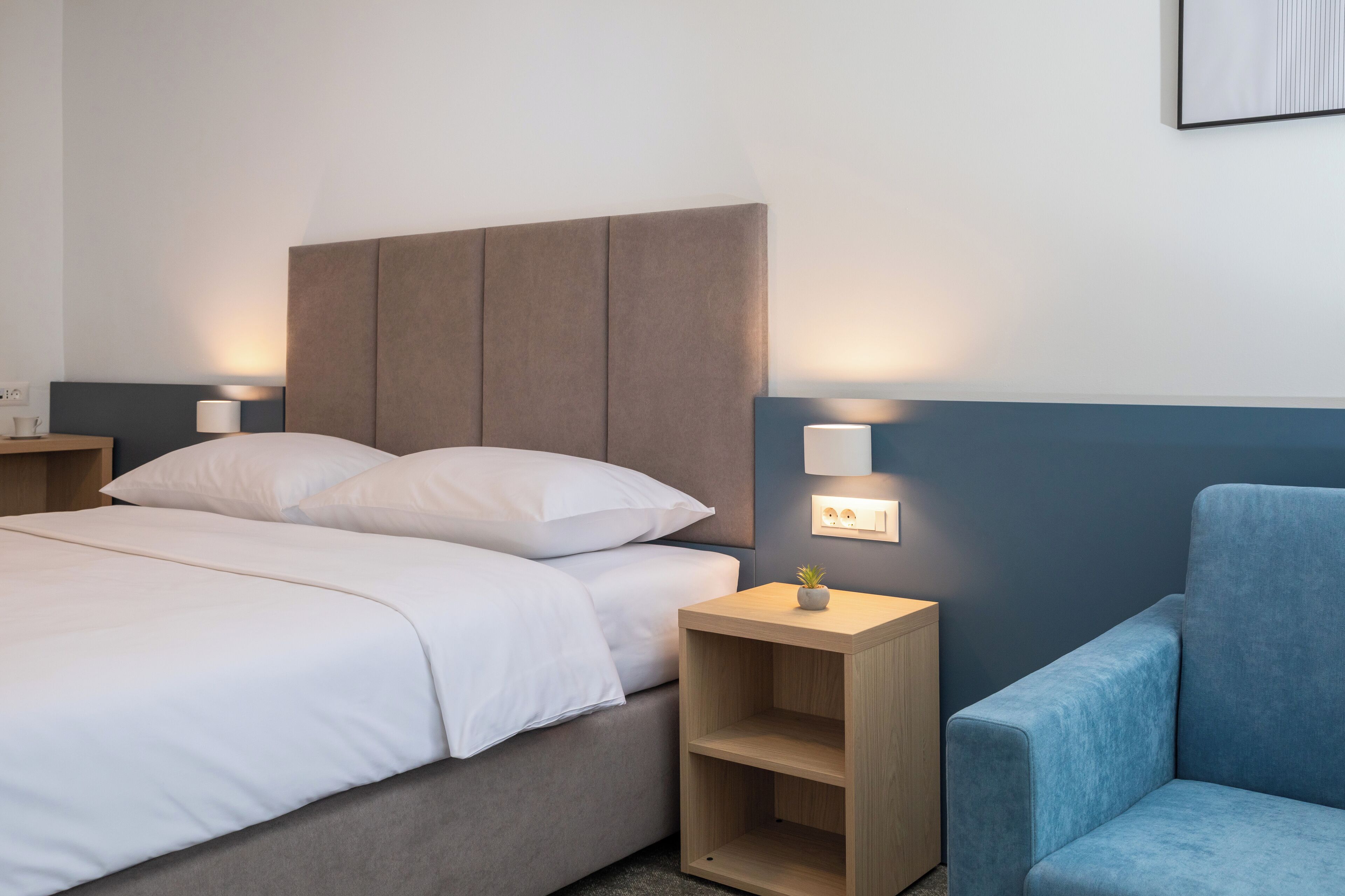 A modern hotel room with a bed, bedside tables, a nightstand, and a blue armchair.