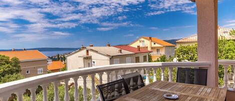A balcony with a wooden table and chairs, a white balustrade, and a view of houses and a body of water.
