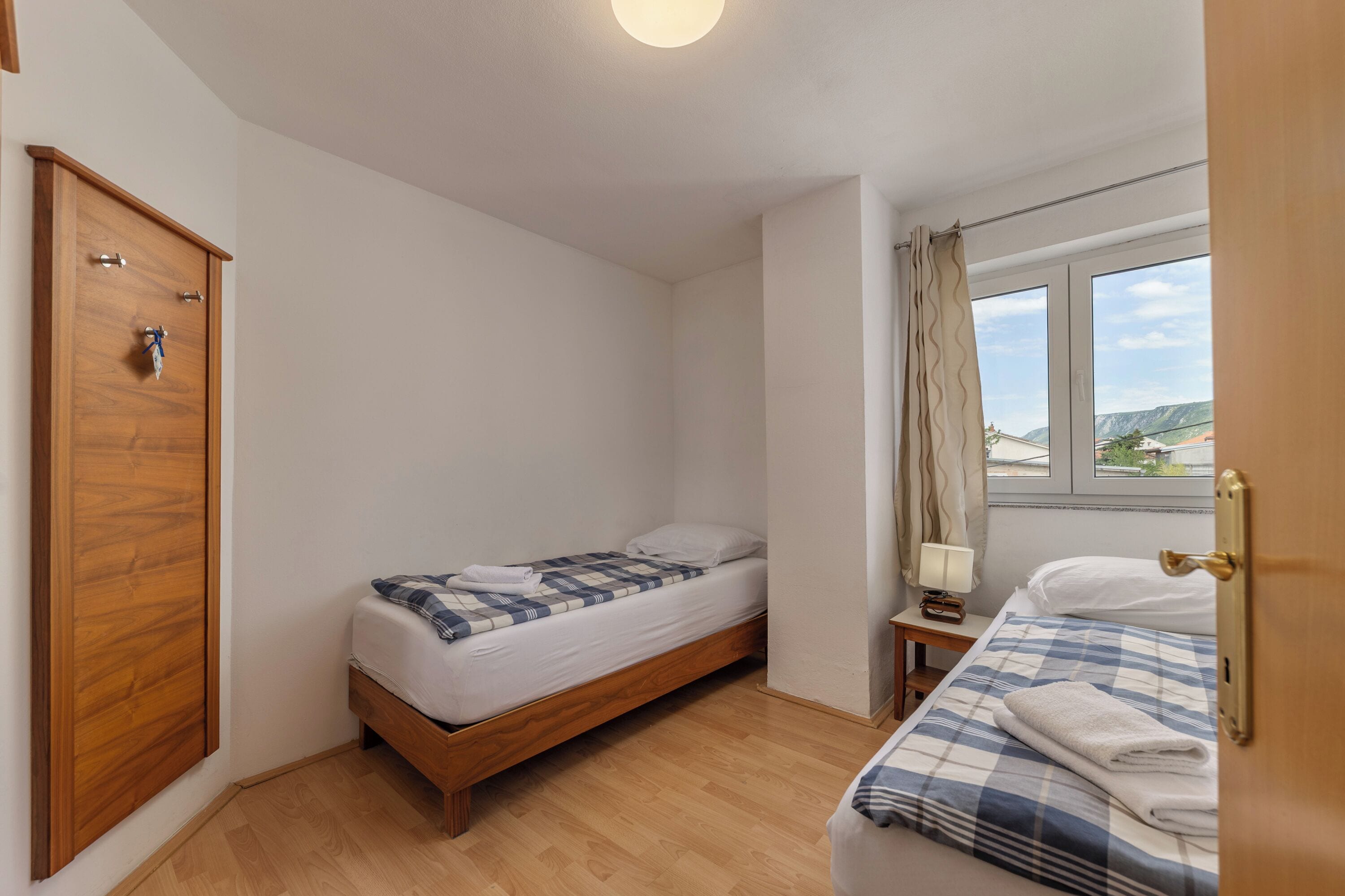 A bedroom with a wooden wardrobe, a single bed with a checkered blanket, a bedside table with a lamp, and a window with curtains.