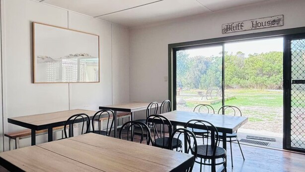 A dining area with tables and chairs, a large window with a view of greenery, and a sign above the window reading "Bluff House".