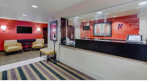 Un espace d’accueil d’hôtel avec un mur rouge, un comptoir noir, un téléviseur à écran plat et un coin salon composé de deux chaises et d’un banc.