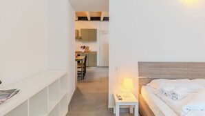 Ein Hotelzimmer mit einem Bett, einem Nachttisch mit Lampe, einem kleinen Tisch und Blick in den Küchenbereich.