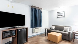 A modern living room with a flat-screen TV, a built-in mini-fridge, a microwave, a gray sofa, a wooden coffee table, and a blue curtain. - Americas Best Value Inn Searcy (Searcy)