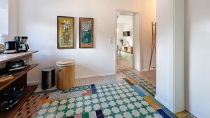 Un pasillo con suelo de baldosas, una cafetera, un cubo de basura y dos obras de arte enmarcadas en la pared.