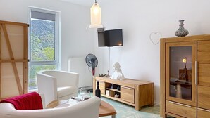 A living room with a white sofa, a wooden cabinet, a TV mounted on the wall, and a large window with a view of trees. - Apartmenthaus 110 by Interhome (Sankt Goar)