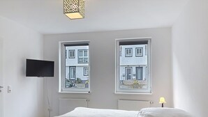 A bedroom with a bed, a flat-screen TV mounted on the wall, two windows with views of buildings, and a radiator. - Apartmenthaus 110 by Interhome (Sankt Goar)