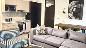 A modern living room with a grey sofa, striped armchair, and round coffee table. A kitchen is visible in the background with a microwave and cabinets. - The Henlee Apartment Sandton (Sandton)