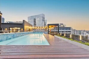 Una piscina en la azotea con cubierta de madera, rodeada de edificios modernos y luces colgantes.
